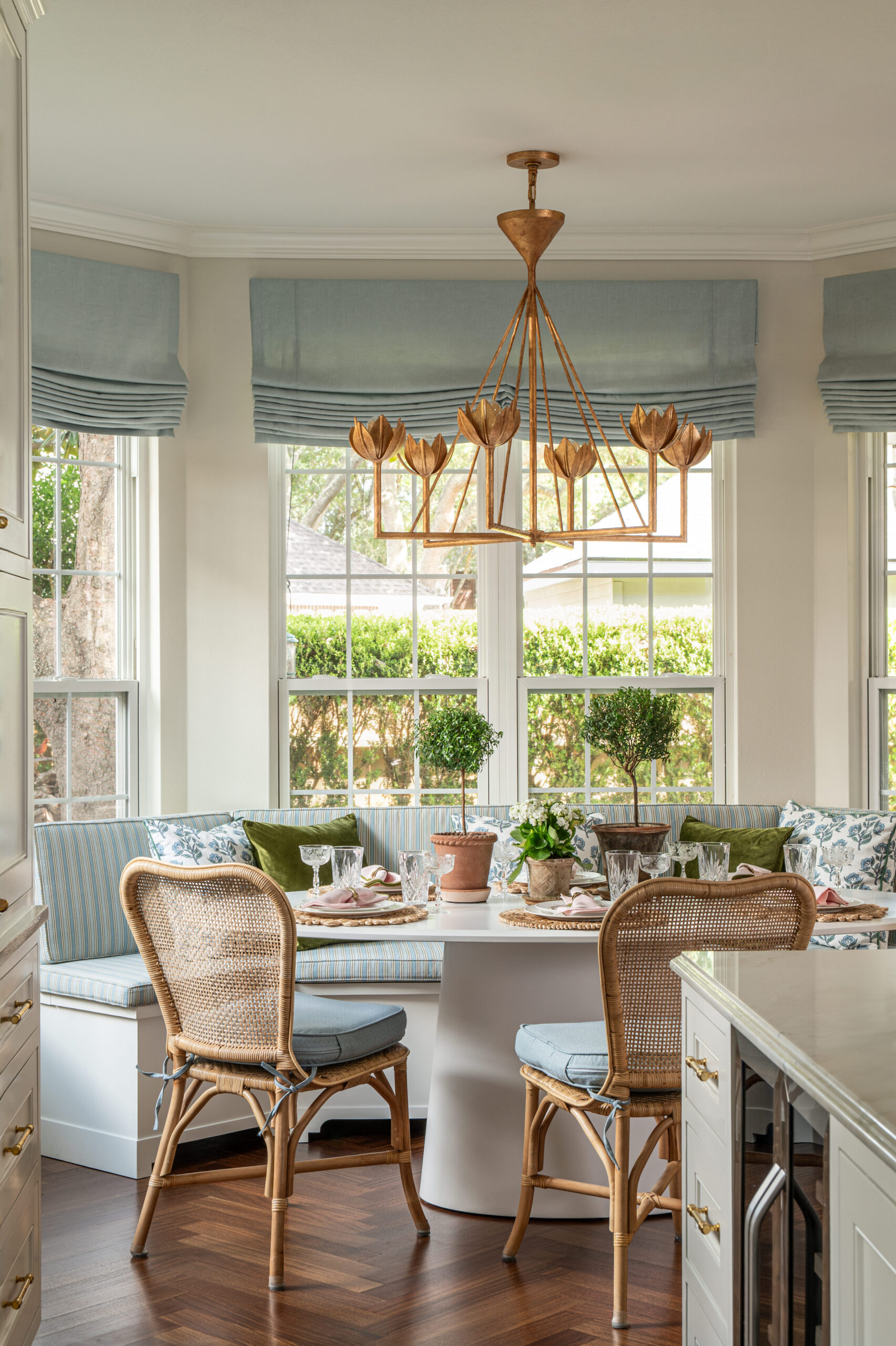 Bright breakfast nook surrounded by windows, complete with a custom banquette, rattan chairs, and a gold petal chandelier above the round table.