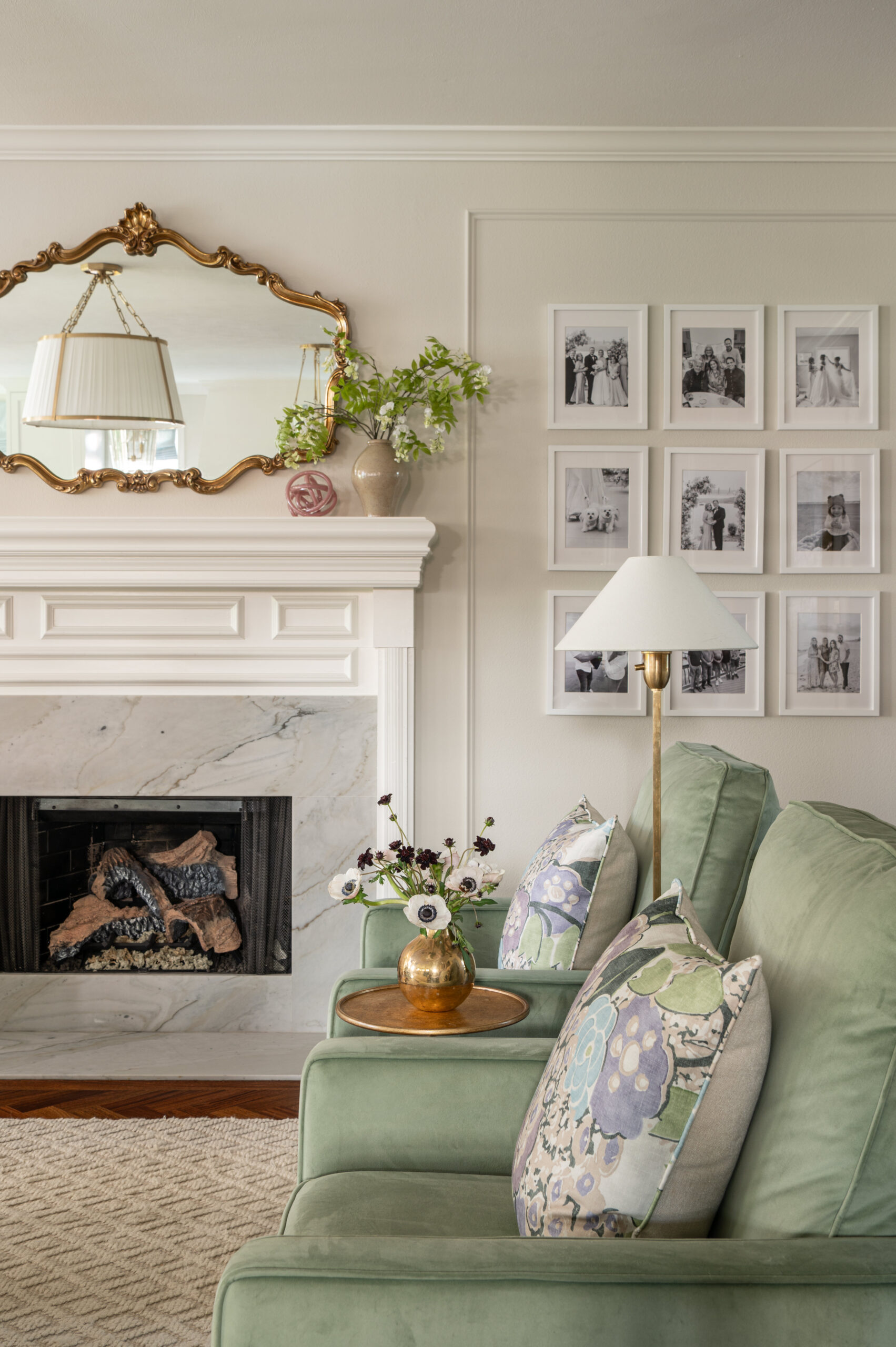 Green armchairs with floral pillows sit in front of a traditional fireplace, with a vintage gold mirror and black-and-white photo grid adding personal warmth.